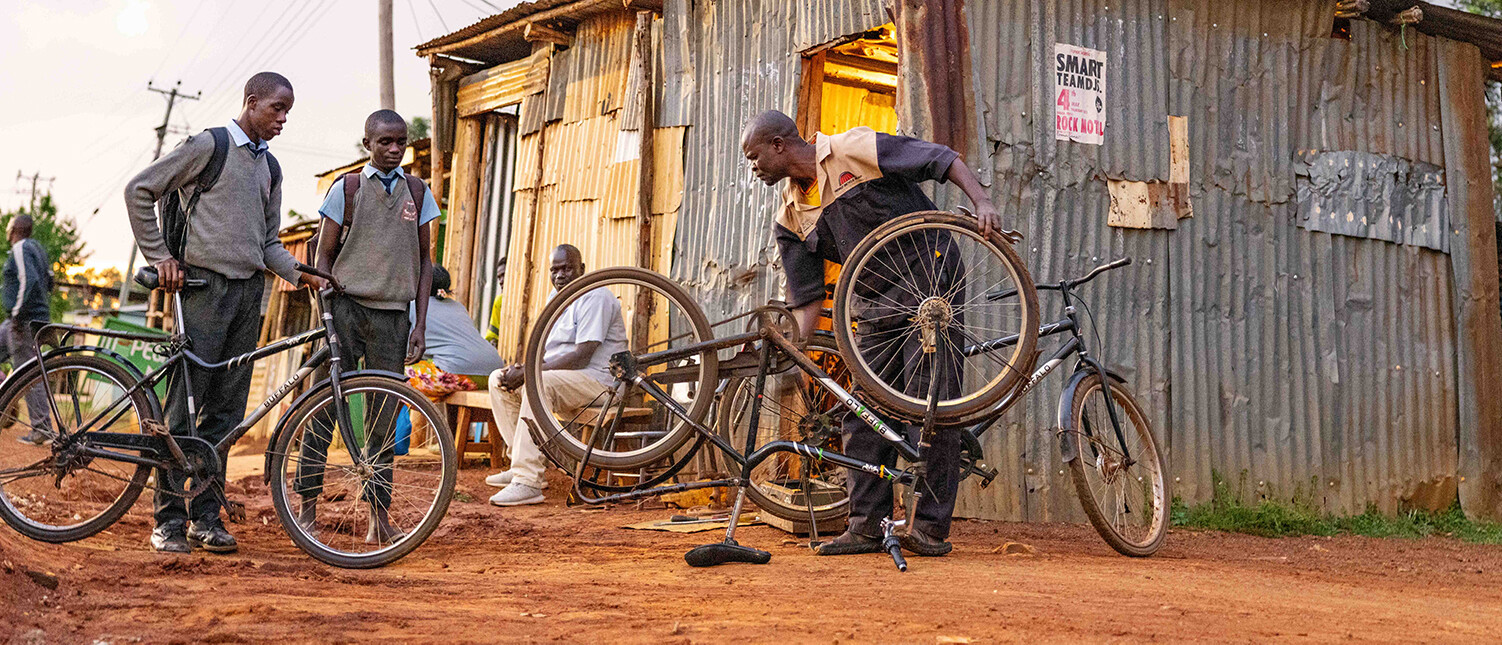 Plusieurs personnes sont rassemblées devant une petite boutique en tôle ondulée, un homme réparant un vélo tandis que deux élèves en uniforme observent.