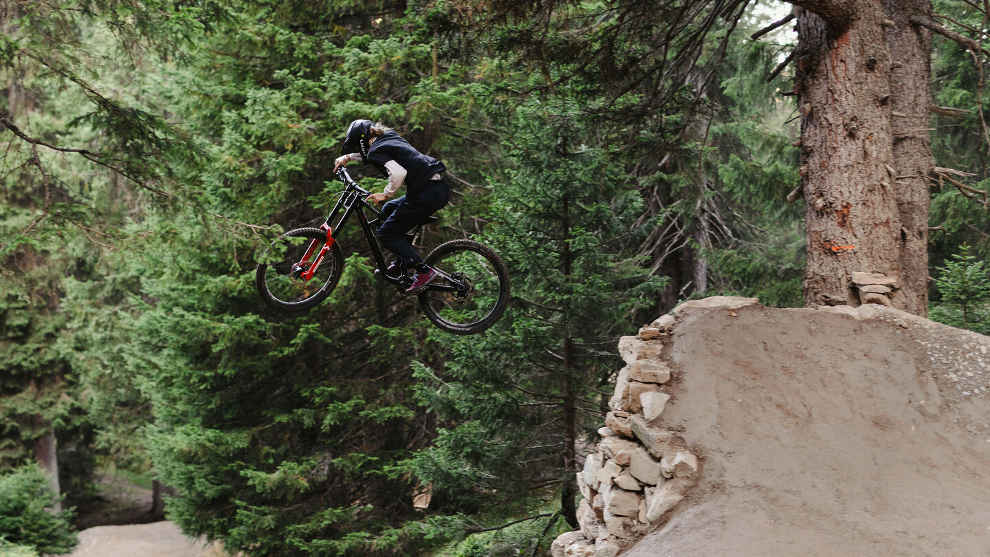 A mountain biker jumps off a ramp with his bike in a dense forest. The rider is wearing a helmet and dark clothing, flying over a massive dirt jump.