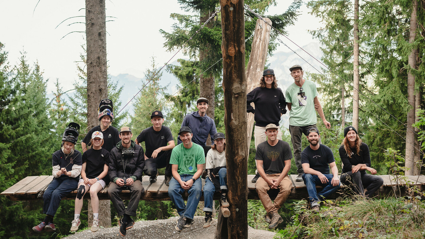 A group of mountain bikers and crew members posing together on a wooden platform in the forest. The individuals are wearing sporty clothes and helmets, smiling at the camera.