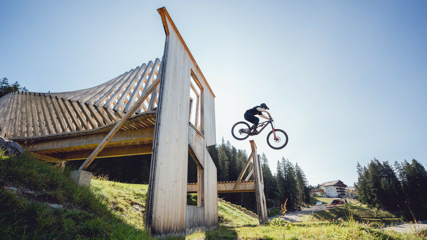 Een mountainbiker springt over een houten ramp in een bikepark. De massieve houten constructie steekt af tegen de heldere lucht en de omliggende natuur.