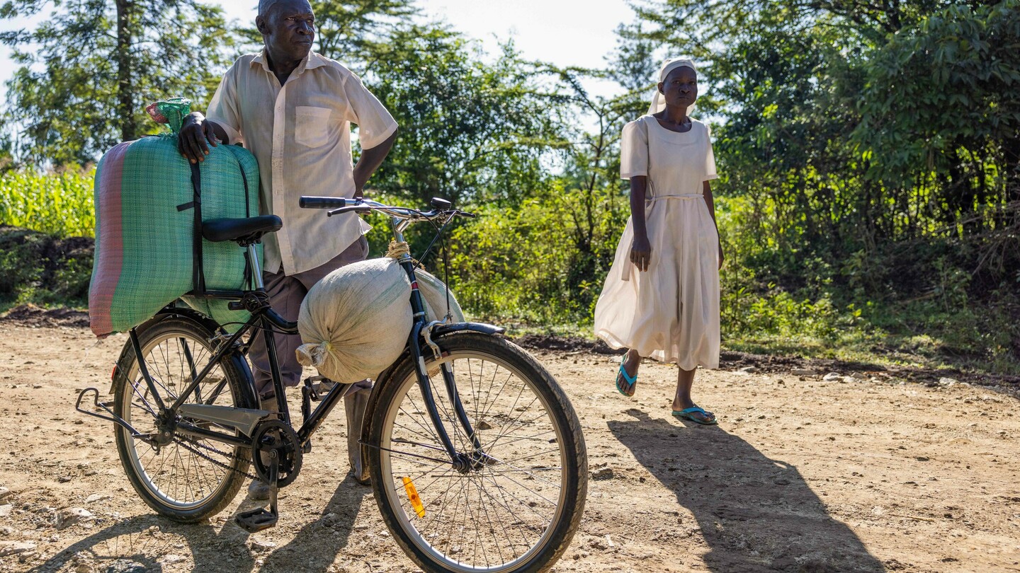 Un homme se tient à côté d'un vélo chargé sur un chemin rural, tandis qu'une femme en robe claire passe à côté. Une verdure luxuriante est visible à l'arrière-plan.