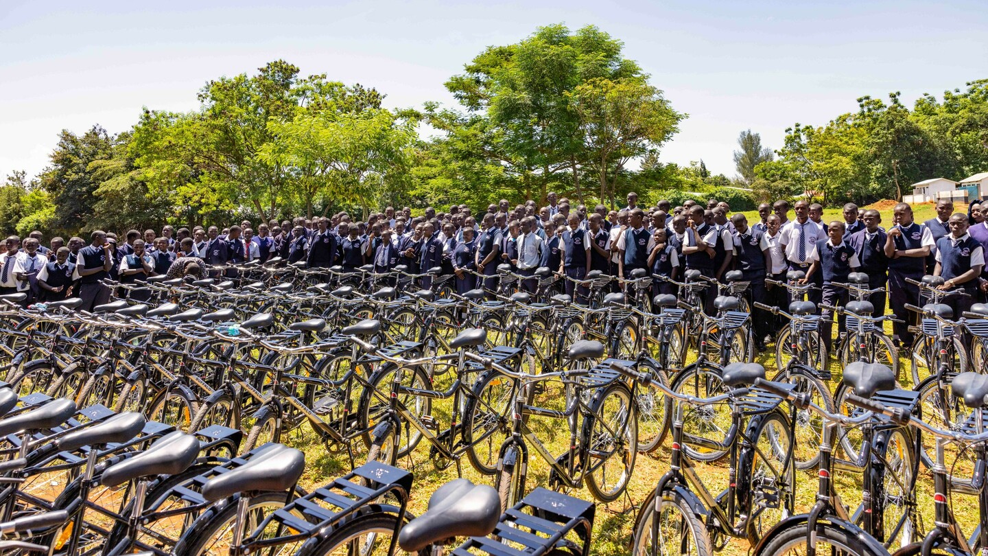 Een grote groep leerlingen in uniformen staat achter een rij gestapelde fietsen in de open lucht. Op de achtergrond zijn groene bomen en een heldere lucht te zien.