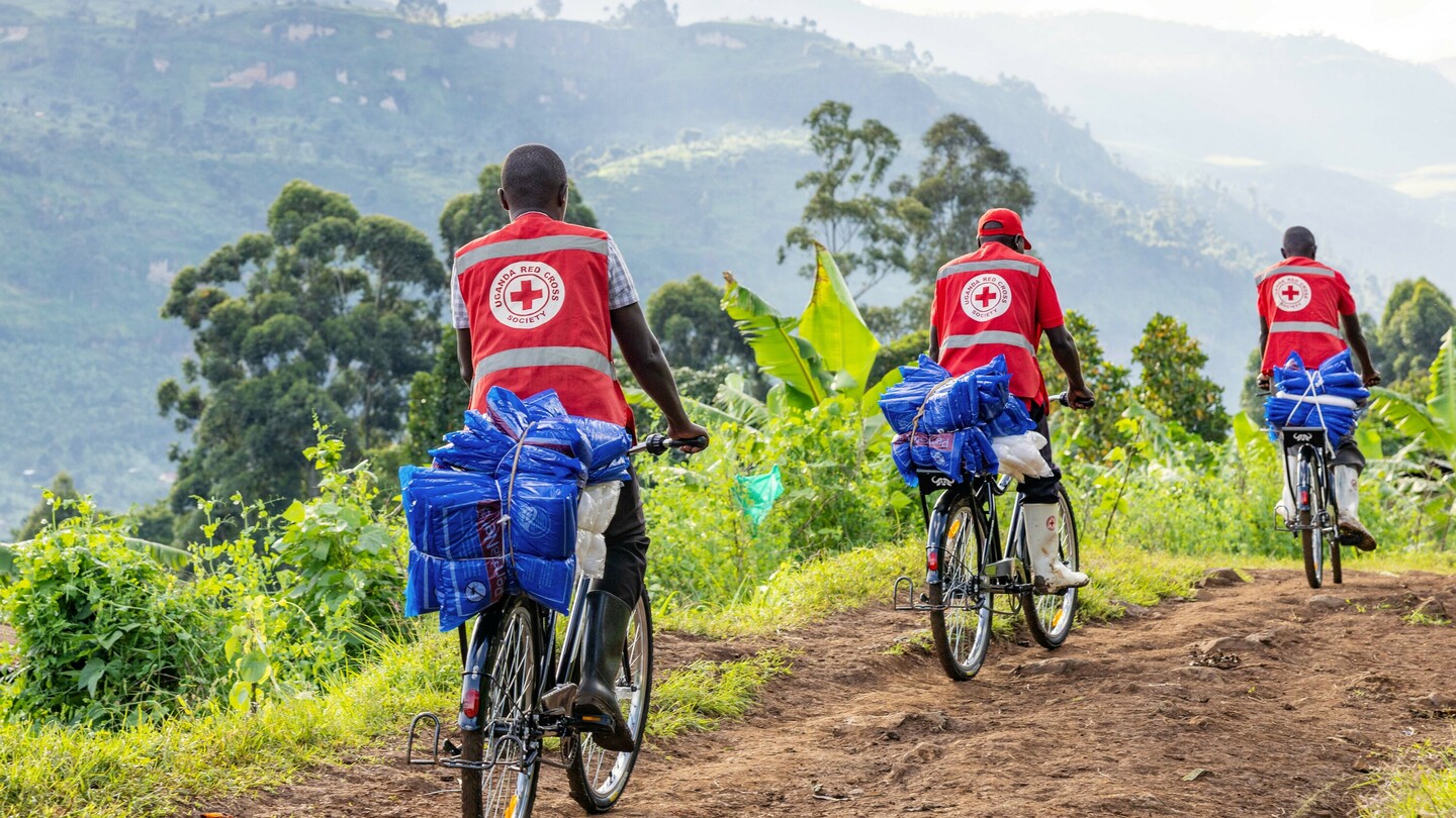 Drie mannen in rode vesten met het logo van Uganda Red Cross Society fietsen met beladen fietsen met blauwe pakketten op een landelijk pad door groen landschap.