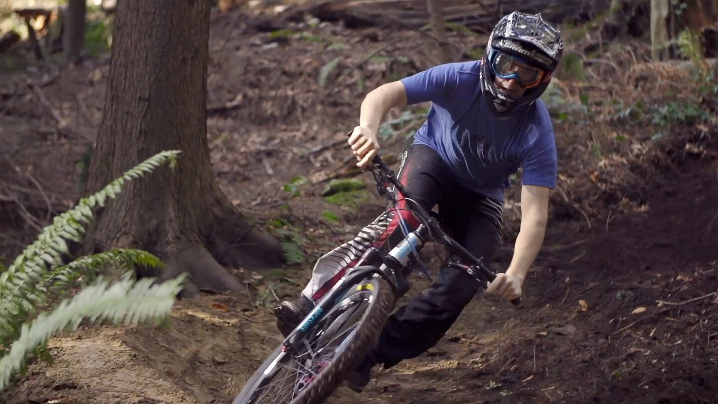 A mountain biker wearing a helmet and goggles riding through a turn in a forest. The rider is dressed in a blue T-shirt and black pants, skillfully maneuvering the bike on the uneven, dirt terrain.