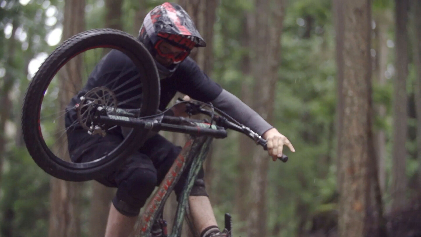 Mountainbiker met helm en bril voert een sprong of trick uit in een bos, met het voorwiel omhoog.