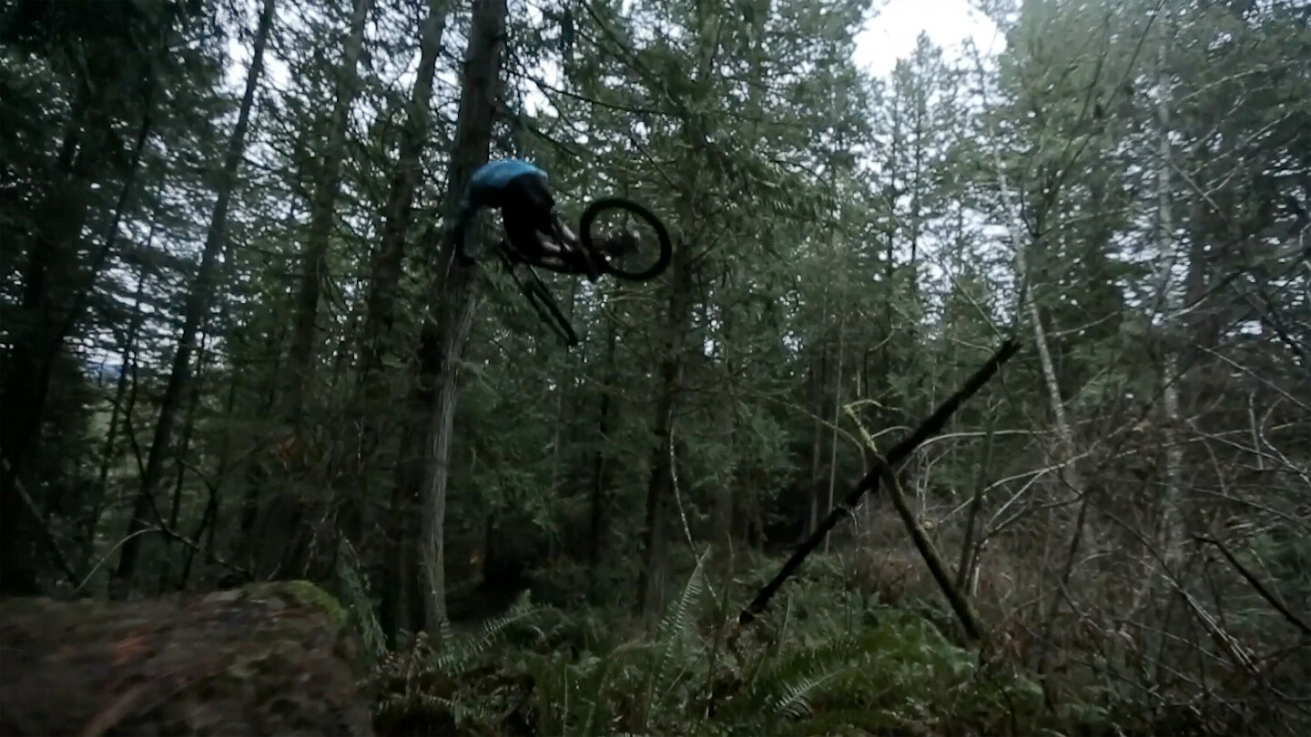 Een mountainbiker in de lucht tijdens een sprong in een dicht bosgebied. De rijder draagt donkere kleding en het bos is gevuld met hoge bomen en dicht struikgewas.