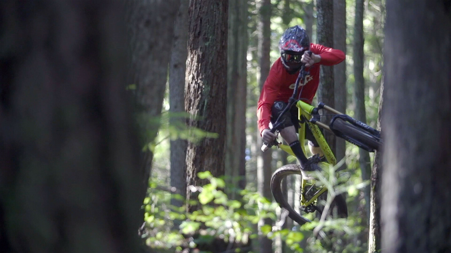 Vététiste portant un casque et un maillot rouge effectuant un saut dans une forêt dense, avec le vélo incliné sur le côté.