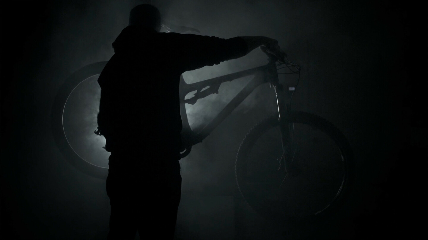 Silhouet van een persoon die een mountainbike vasthoudt in een donkere, rokerige omgeving met licht dat van achteren schijnt.