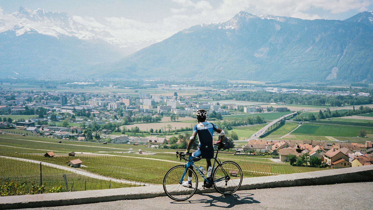 Un cycliste se tient à côté de son vélo et contemple une vallée avec une ville, des champs et des montagnes en arrière-plan par une journée ensoleillée.