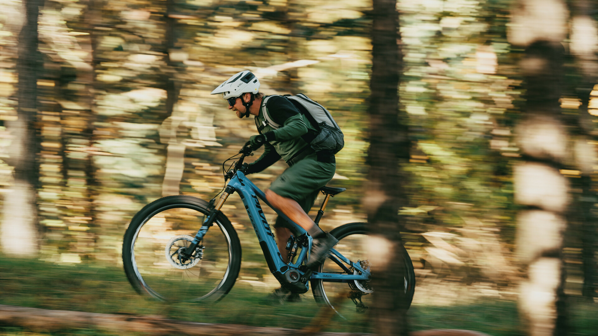 Mountainbiker met helm en rugzak rijdt met hoge snelheid door een bos op een blauwe fiets.